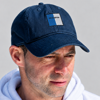 A man wearing a navy blue cotton baseball cap with a small rectangular patch on the front that reads 'AN CHLOE', paired with a white hoodie against a light gray background.