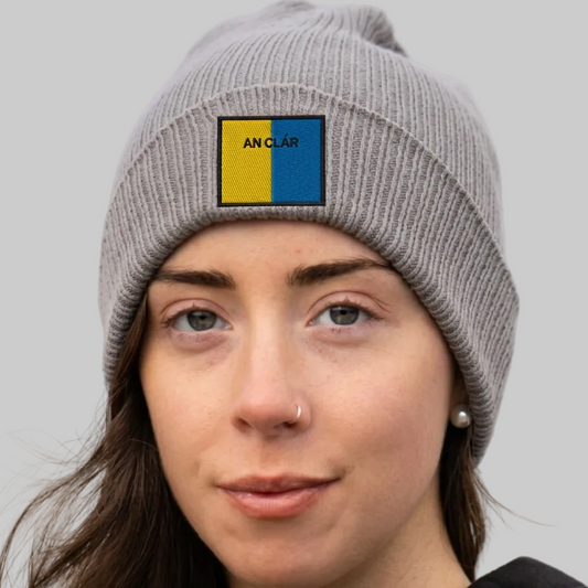 A close-up of a woman wearing a gray ribbed knit beanie with a square patch on the front that reads 'AN CLÁR' in black text over a yellow and blue background. The beanie is photographed outdoors with a blurred natural background.