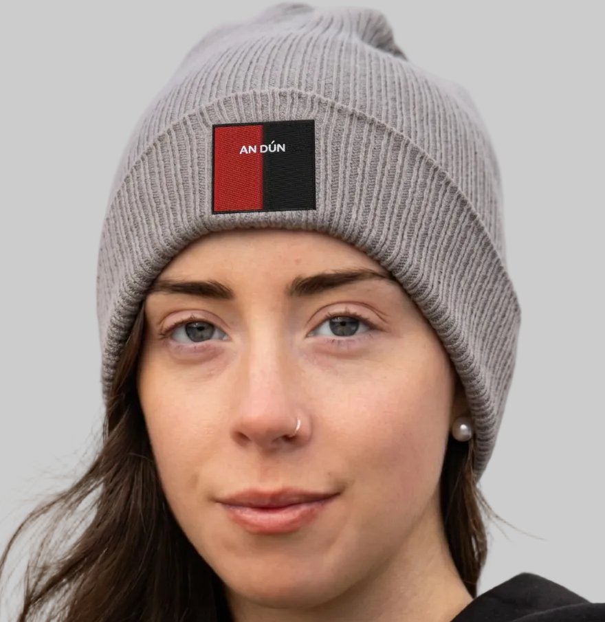 A close-up of a woman wearing a gray ribbed knit beanie with a rectangular front patch that reads 'AN DÜN' in white text over a red and black background. The photo is taken outdoors with a blurred natural background.