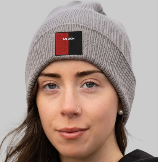 A close-up of a woman wearing a gray ribbed knit beanie with a rectangular front patch that reads 'AN DÜN' in white text over a red and black background. The photo is taken outdoors with a blurred natural background.