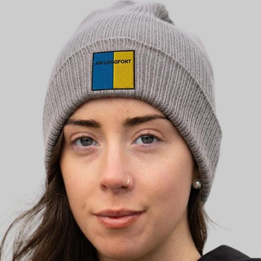 A close-up of a woman wearing a gray ribbed knit beanie with a square embroidered patch that reads 'AN LONGFORT' in black text over blue and yellow blocks. The background is a soft-focus outdoor scene with muted blue-gray tones.