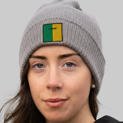 A close-up of a woman wearing a gray ribbed knit beanie with a square patch on the front. The patch is split vertically into green on the left and yellow on the right, with the word 'AN MHÍ ' printed in black on the green section.