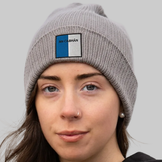 A close-up of a woman wearing a gray ribbed knit beanie with a rectangular embroidered patch on the front. The patch is split vertically into blue and white with the text 'AN CABHÁN in black.