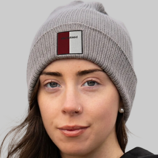 A close-up of a woman wearing a gray ribbed knit beanie with a rectangular front patch that reads 'AN IARMHI’ on a red and white background. The photo is taken outdoors with a softly blurred natural background.