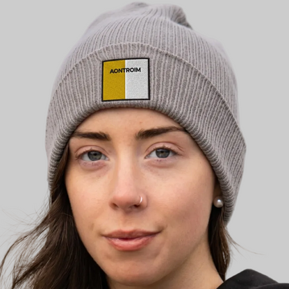 A woman wearing a gray ribbed knit beanie with a square patch that reads 'AONTROIM' in black text over yellow and white blocks. The background is grey.