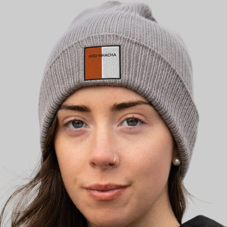 A close-up of a woman wearing a gray ribbed knit beanie with a square patch on the front that reads 'ARD MHACHA' in black text over orange and white blocks. The background is grey.