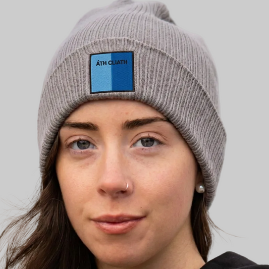 A close-up of a woman wearing a gray ribbed knit beanie with a blue and black square patch on the front that reads ÁTH CLIATH The background is grey.