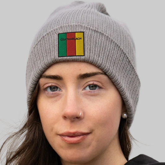 A close-up of a woman wearing a gray ribbed knit beanie with a square patch featuring green, yellow, and red vertical stripes and the word 'Ceatharlach'.