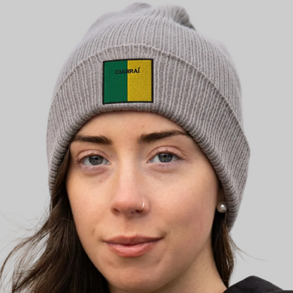 A close-up of a woman wearing a gray ribbed knit beanie with a square patch on the front. The patch is split vertically into green and yellow with the text 'CIARRAÍ at the top.
