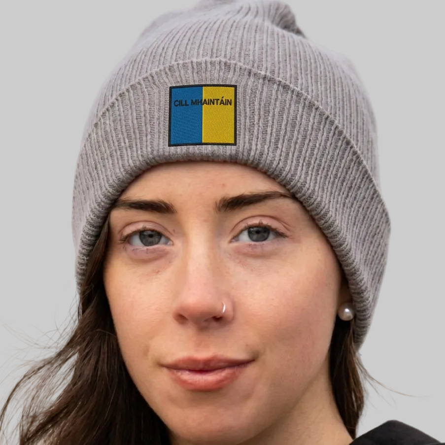 A close-up of a woman wearing a gray ribbed knit beanie with a square patch on the front. The patch is split vertically into blue on the left and yellow on the right, with the text 'CILL MHAINTÁIN' printed in black above the split.