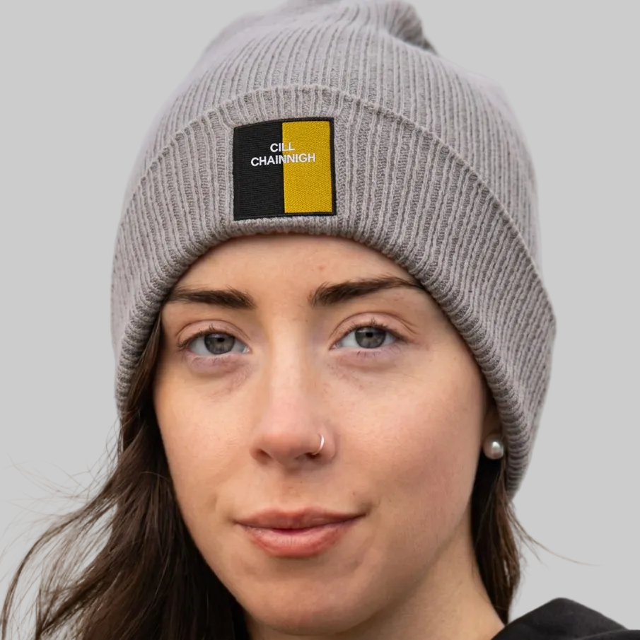 A close-up of a woman wearing a gray ribbed knit beanie with a square black and yellow patch on the front that reads 'CILL CHAINNIGH'. The background is grey.