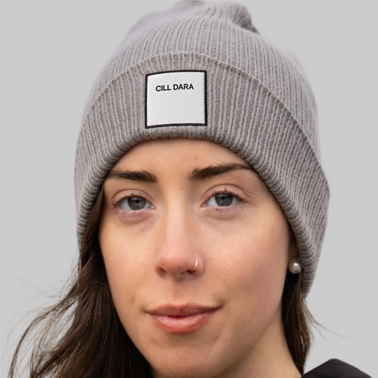 A close-up of a woman wearing a gray ribbed knit beanie with a white rectangular patch that reads 'CILL DARA'. The background is a soft, blurred outdoor scene.