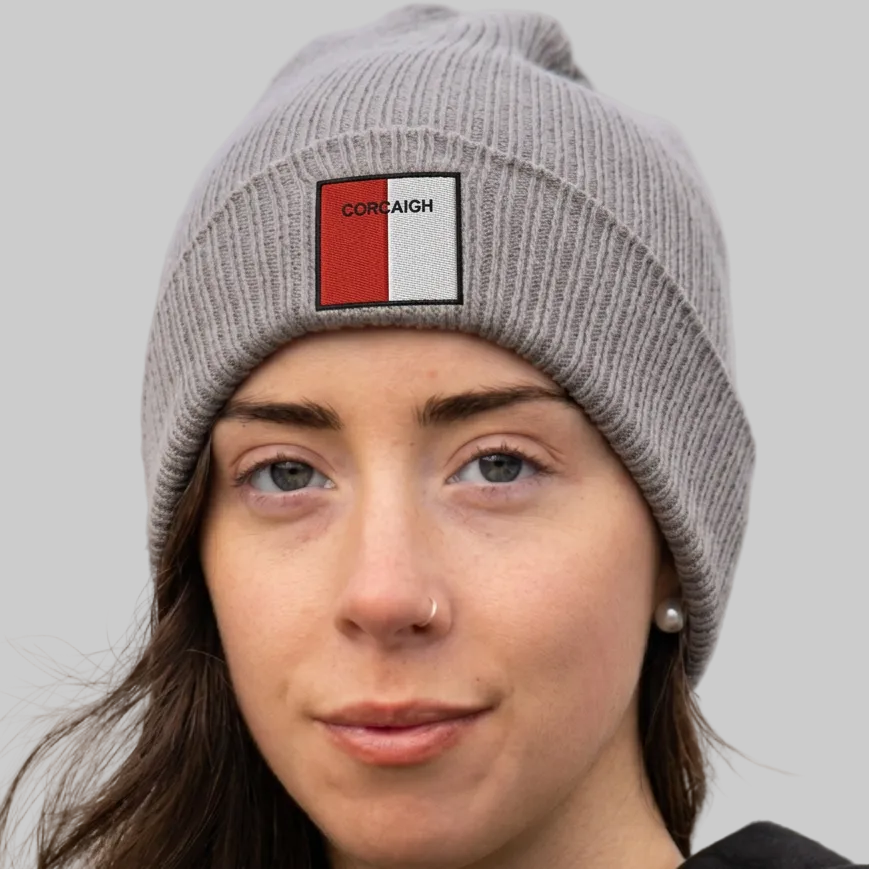 A close-up of a woman wearing a gray ribbed knit beanie with a rectangular patch on the front. The patch is split vertically into red and white with the word 'CORCAIGH' printed in black.