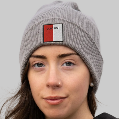 A close-up of a woman wearing a gray ribbed knit beanie with a rectangular patch on the front. The patch is split vertically into red and white with the word 'CORCAIGH' printed in black.