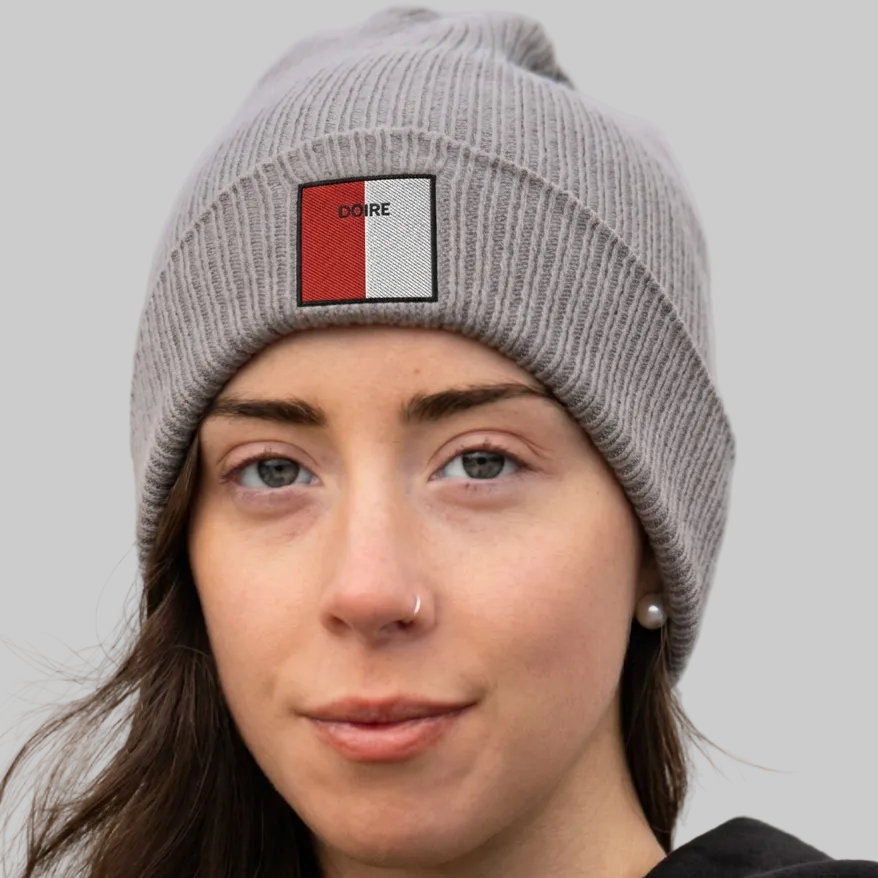 A close-up of a woman wearing a gray ribbed knit beanie with a square patch on the front. The patch is split vertically into red and white on the left and black and white on the right, with the word 'DOIRE' printed in black on the red section.