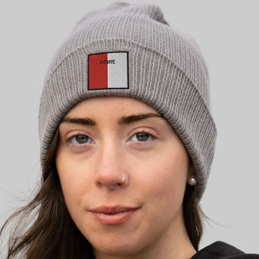 A close-up of a woman wearing a gray ribbed knit beanie with a square patch on the front. The patch is split vertically into red and white on the left and black and white on the right, with the word 'DOIRE' printed in black on the red section.