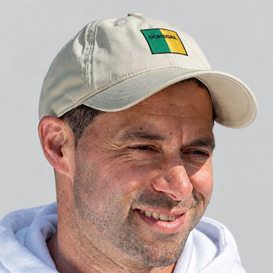 A man wearing a light beige baseball cap with a blue and yellow rectangular patch on the front, photographed against a plain light gray background.