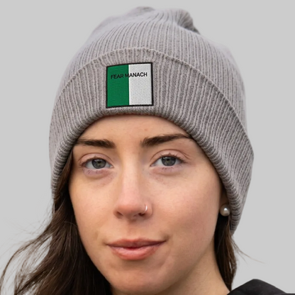 A close-up of a woman wearing a gray ribbed knit beanie with a square patch on the front. The patch is split vertically into green and white with the text 'FEAR MANACH' printed in black.