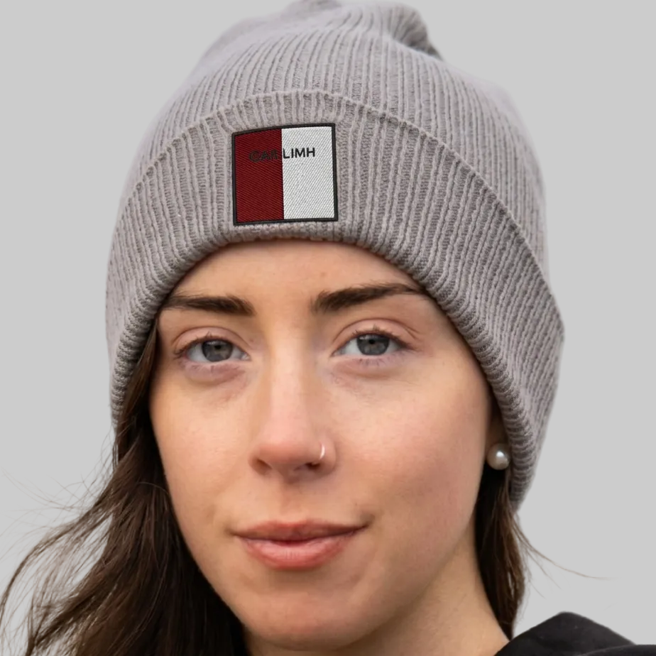 A close-up of a woman wearing a gray ribbed knit beanie with a square patch on the front. The patch is split vertically into red and white with the text 'GAILLIMH' printed on it.
