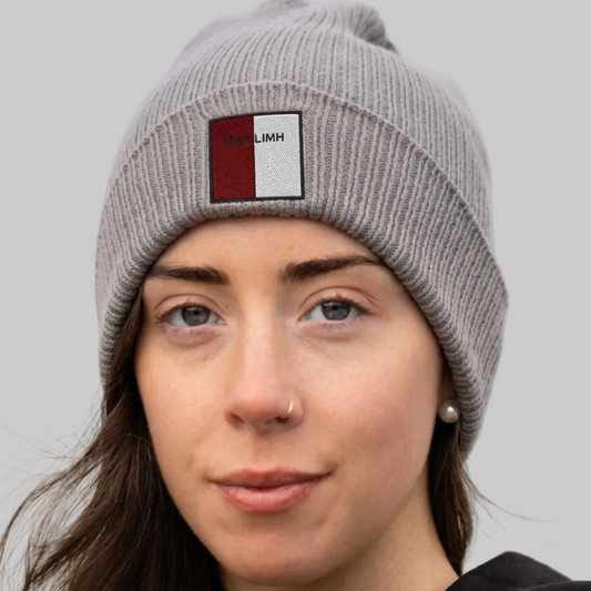 A close-up of a woman wearing a gray ribbed knit beanie with a square patch on the front. The patch is split vertically into red and white with the text 'GAILLIMH' printed on it.