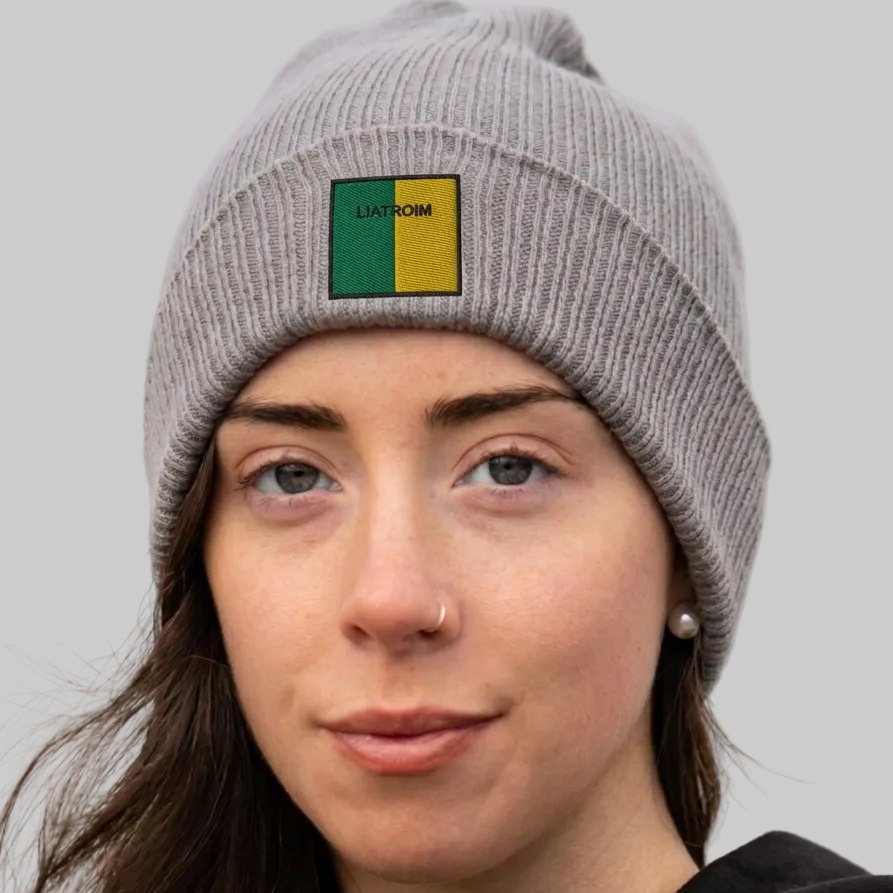 A close-up of a woman wearing a gray ribbed knit beanie with a square patch on the front. The patch is split vertically into green and yellow with the text 'LIATROIM' printed on it.
