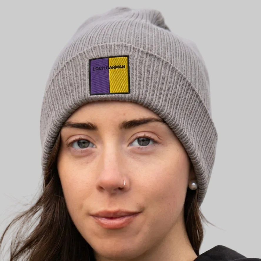 A woman wearing a gray ribbed knit beanie with a square patch that reads 'LOCH GARMAN' in black text over purple and yellow block.
