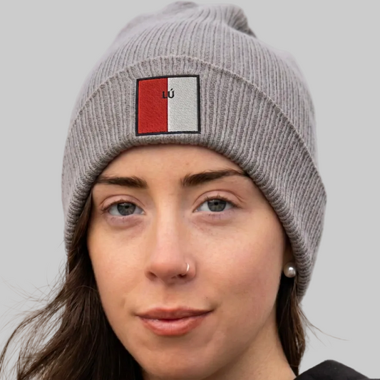 A close-up of a woman wearing a gray ribbed knit beanie with a square embroidered patch on the front. The patch is split vertically into red and white with the letters 'Lú’ in the center.