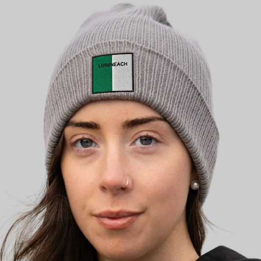 A woman wearing a gray ribbed knit beanie hat with a square patch on the front featuring green, white, and black, LUIMNEACH.