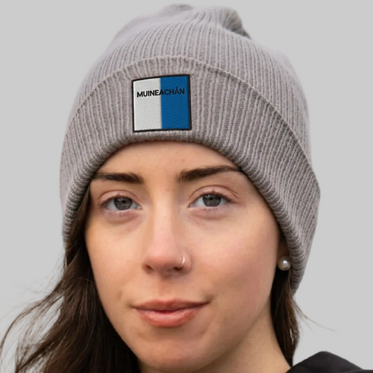 A close-up of a woman wearing a gray ribbed knit beanie with a square patch on the front. The patch is white on the left and blue on the right, with the word 'MUNEACHÁN' printed in black on the white section.