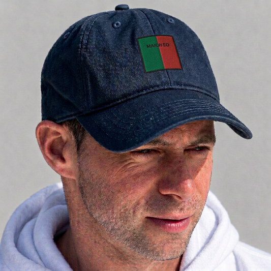 A man wearing a dark navy blue baseball cap with a small rectangular patch on the front that reads 'MADE' in black text over green and red blocks. The man is also wearing a white hoodie against a plain light gray background.