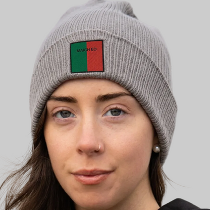 A close-up of a woman wearing a gray ribbed knit beanie with a square patch on the front. The patch is split vertically into green on the left and red on the right, with the text 'MAIGH EO' printed in black on the green section.