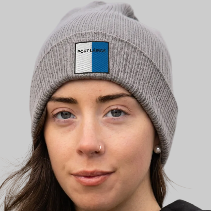 A close-up of a woman wearing a gray ribbed knit beanie with a square PORT LÁIRGE logo patch on the front, set against a softly blurred outdoor background.