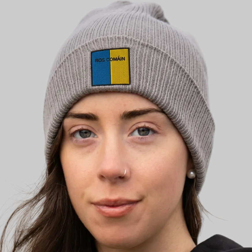 A close-up of a woman wearing a gray ribbed knit beanie with a square patch on the front. The patch is split vertically into blue and yellow, with the text 'ROS COMÁIN' printed in black.
