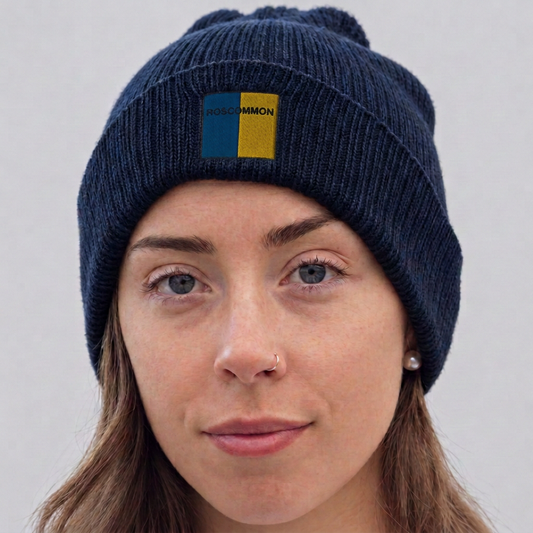 A close-up of a woman wearing a navy blue ribbed knit beanie with a pom-pom. The beanie features a front patch with a vertical split design of blue and yellow, with the brand name 'Roscommon' printed in black on the yellow section.