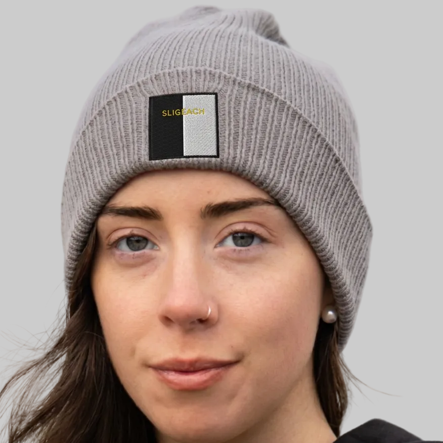 A woman wearing a gray ribbed knit beanie hat with a black and white rectangular patch on the front that reads 'SLIGEACH' in gold.