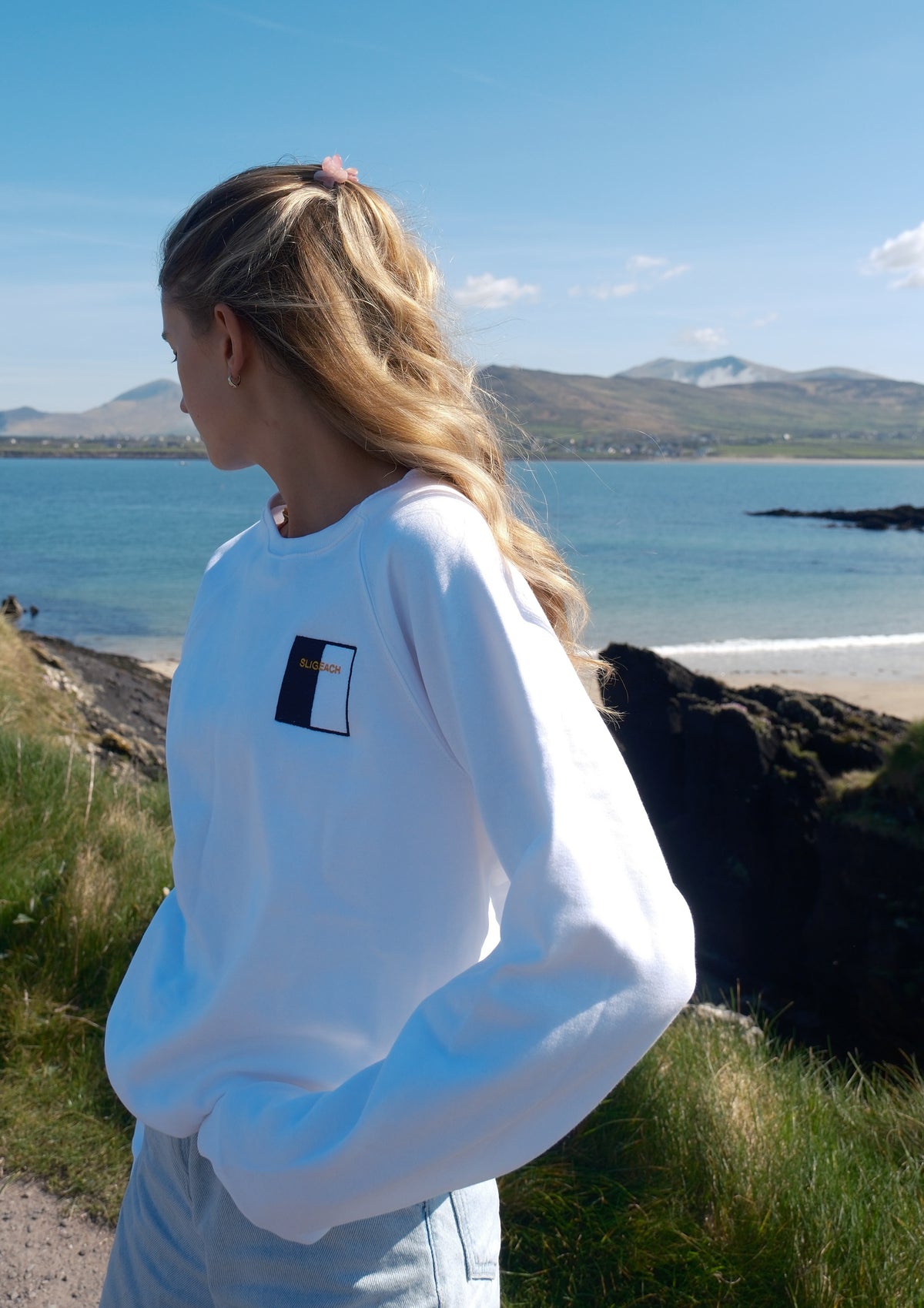 A woman with blonde hair in a ponytail stands outdoors by the sea, wearing a white crewneck sweatshirt with a small black and orange graphic on the left chest.
