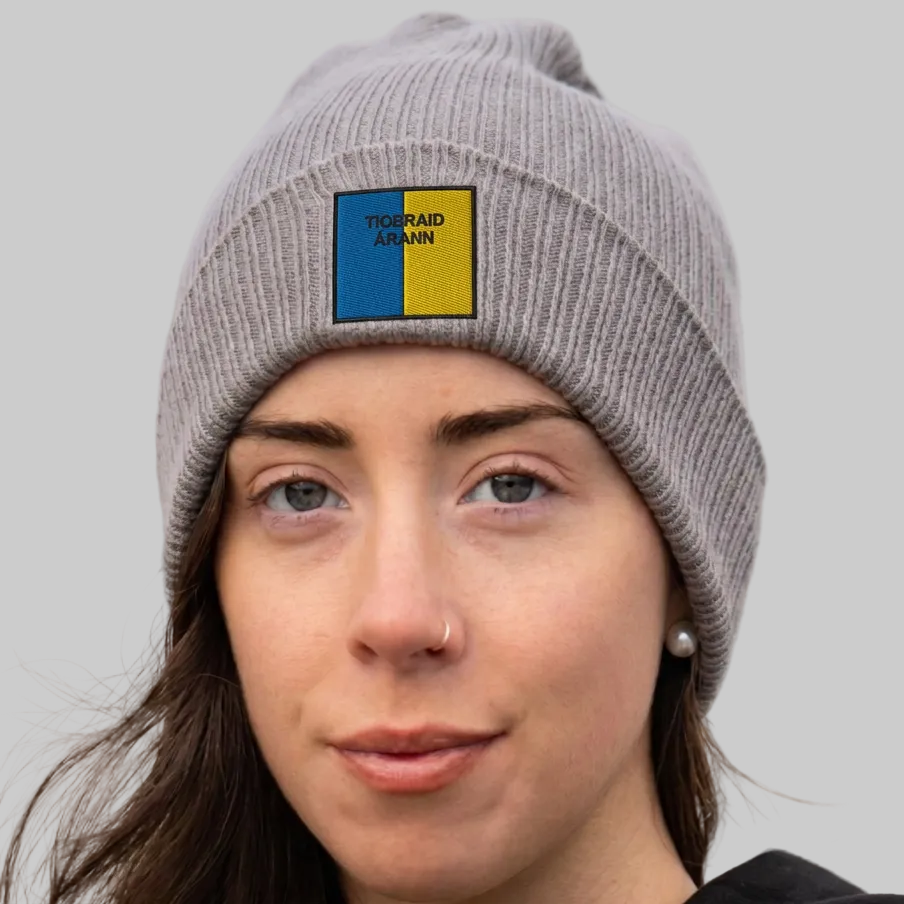 A close-up of a woman wearing a gray ribbed knit beanie with a square patch on the front. The patch is split vertically into blue and yellow with the text 'TIOBRAID ÁRANN’ .