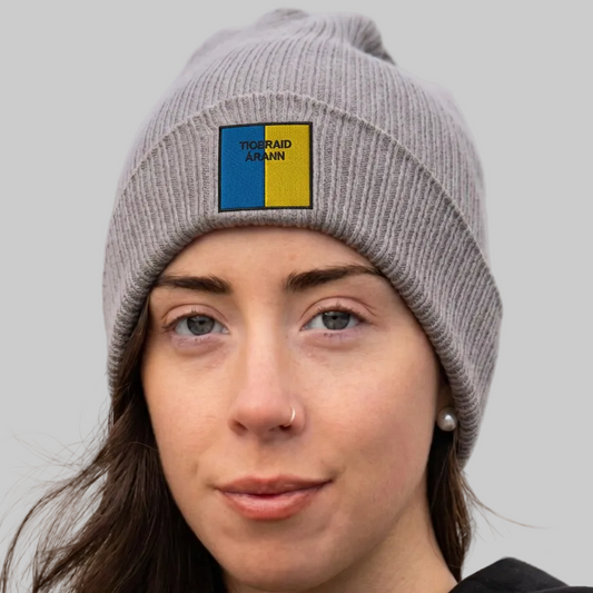 A close-up of a woman wearing a gray ribbed knit beanie with a square patch on the front. The patch is split vertically into blue and yellow with the text 'TIOBRAID ÁRANN’ .