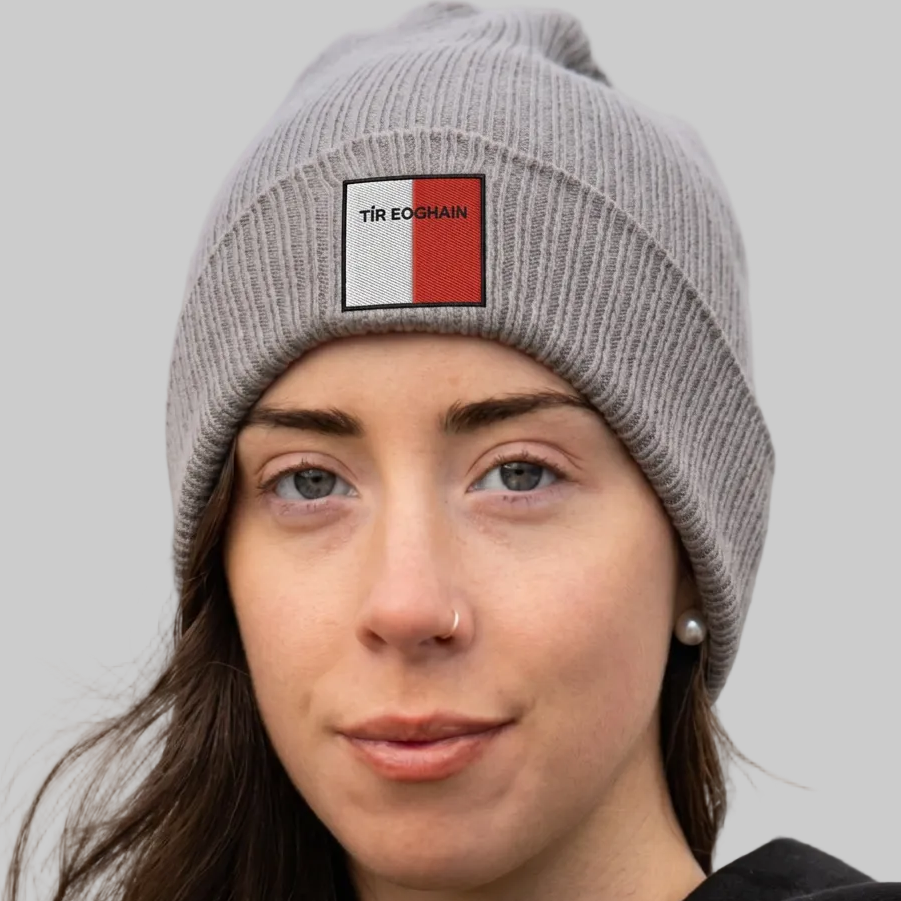 A close-up of a woman wearing a gray ribbed knit beanie with a square embroidered patch on the front. The patch is white on the left side with the black text 'TIR EOGHAIN' and has a red stripe on the right side.