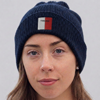 A close-up of a woman wearing a navy blue ribbed knit beanie with a small front patch that reads TYRONE in white text over a red and white square.
