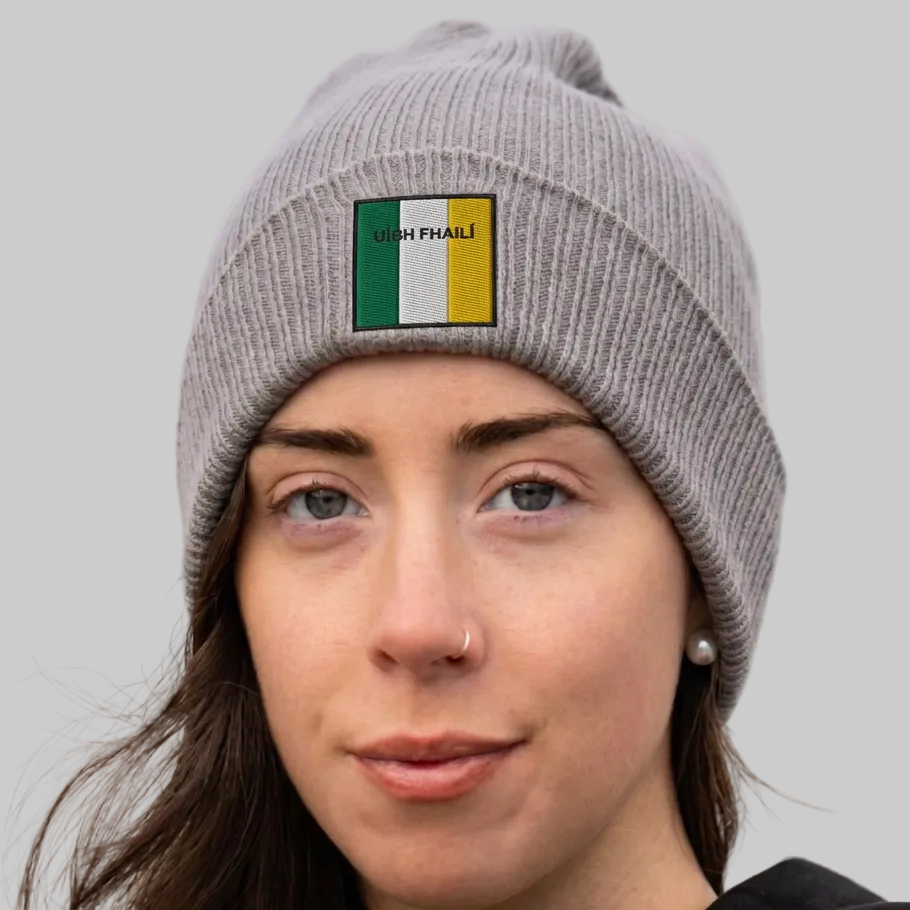 A woman wearing a gray ribbed knit beanie with a small rectangular patch featuring the Irish flag and the text 'UIBH FHAILÍ'. The background is grey.