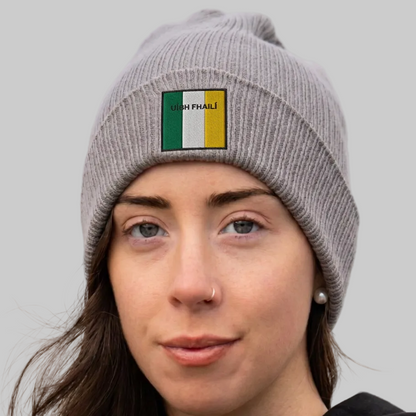 A woman wearing a gray ribbed knit beanie with a small rectangular patch featuring the Irish flag and the text 'UIBH FHAILÍ'. The background is grey.