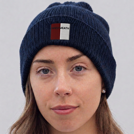 A close-up of a woman wearing a navy blue ribbed knit beanie with a small front patch that reads 'WEST MEATH' in white text over a red and white background.