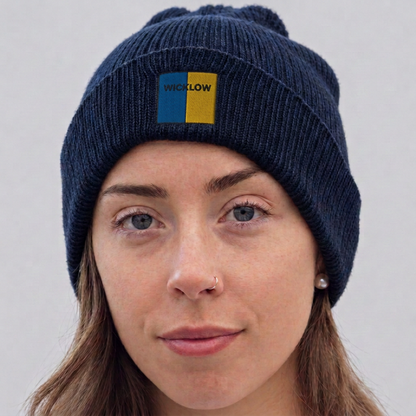 A close-up of a woman wearing a navy blue ribbed knit beanie with a pom-pom. The beanie features a small rectangular patch on the front with the word 'WICKLOW' in yellow text over a split blue and yellow background.